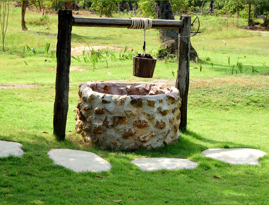 A stone-lined water well sits in a sunny green field with stone pavers leading toward it, symbolizing an adoptee's journey of returning to their original well, their birth parents.